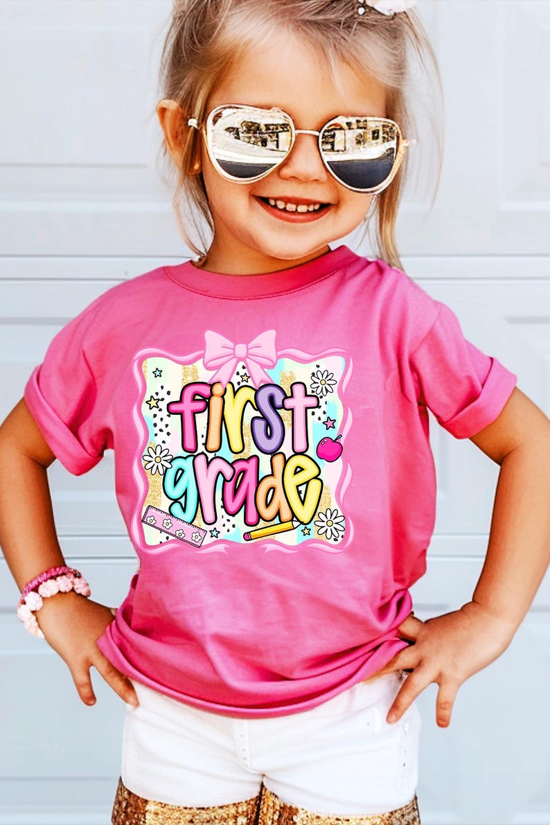 May include: A pink t-shirt with the words "First Grade" in colorful bubble letters. The graphic includes a pink bow, stars, flowers, a pencil, and an apple. The shirt is worn by a child wearing sunglasses and a pink bracelet.