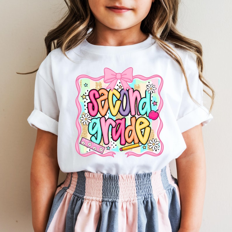May include: A white short-sleeved t-shirt featuring a colorful graphic print that says second grade in bubble letters. The design includes a pink bow, a ruler, a pencil, an apple, and daisies, all framed within a pink scalloped border.