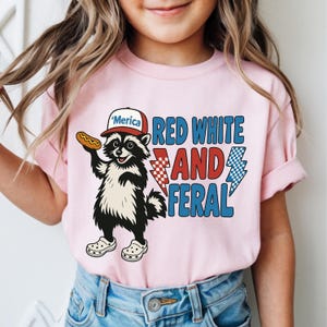 May include: Pink t-shirt featuring a raccoon wearing a hat that says "'Merica" and holding a hot dog. The shirt has the text "RED WHITE AND FERAL" with lightning bolt graphics. The raccoon is wearing white clogs.
