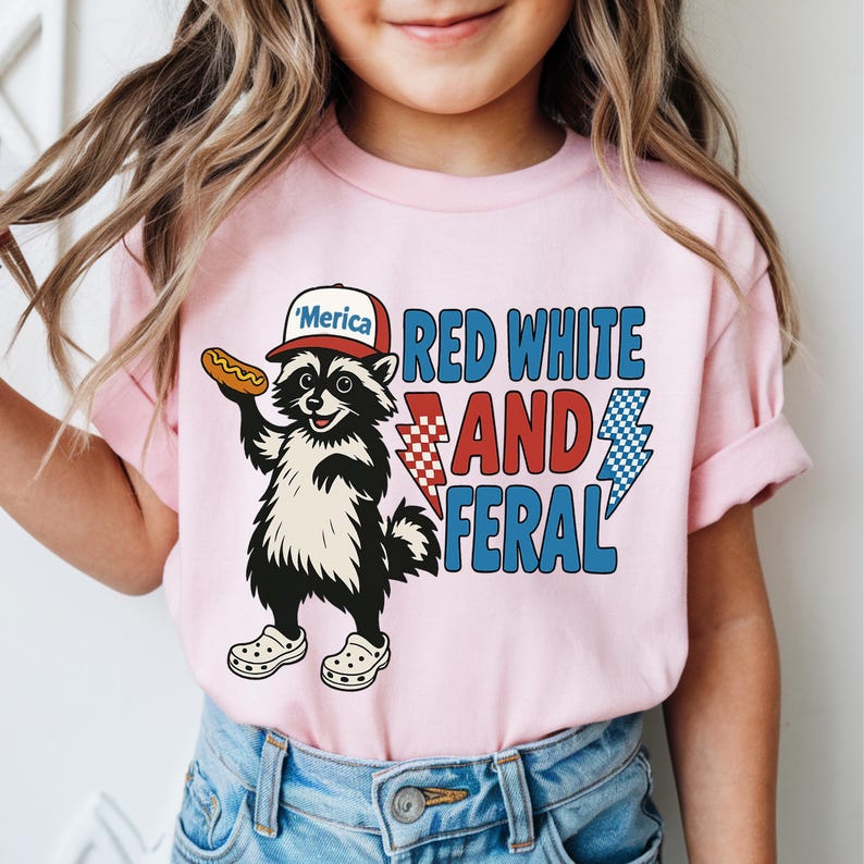 May include: Pink t-shirt featuring a raccoon wearing a hat that says "'Merica" and holding a hot dog. The shirt has the text "RED WHITE AND FERAL" with lightning bolt graphics. The raccoon is wearing white clogs.