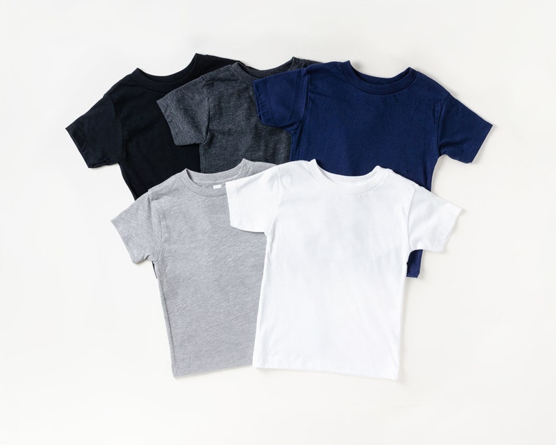 May include: Five short-sleeved t-shirts in various colors: black, charcoal gray, navy blue, light gray, and white. The shirts are arranged in a layered, overlapping pattern, showcasing the different hues and styles.