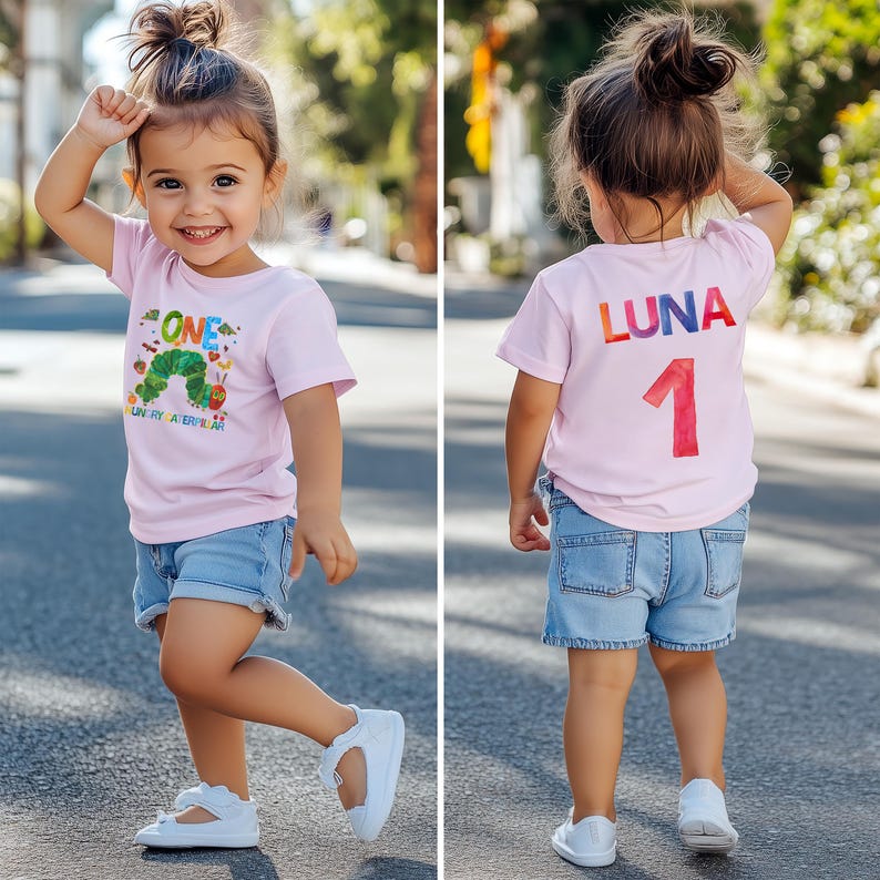 May include: A young child wearing a light pink t-shirt and denim shorts. The front of the shirt features the text "ONE" and a colorful caterpillar design. The back of the shirt displays the name "LUNA" and the number "1". The child is wearing white shoes.