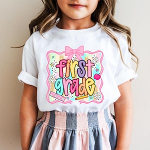 May include: White t-shirt with a colorful "First Grade" graphic. The design includes a pink bow, stars, flowers, a pencil, and an apple. The shirt is worn by a child.