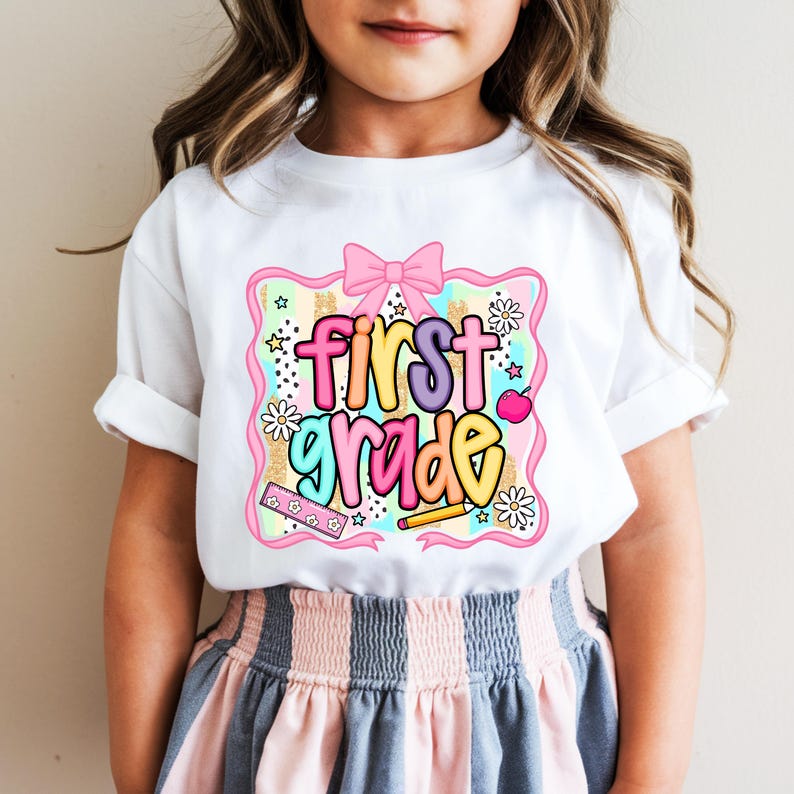 May include: White t-shirt with a colorful "First Grade" graphic. The design includes a pink bow, stars, flowers, a pencil, and an apple. The shirt is worn by a child.