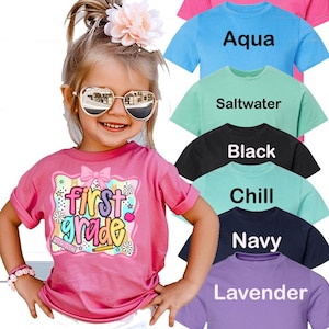 May include: A young girl wearing a pink t-shirt with the words "First Grade" in a colorful design. The image also shows examples of t-shirts in various colors, including hot pink, aqua, saltwater, black, chill, navy, and lavender.