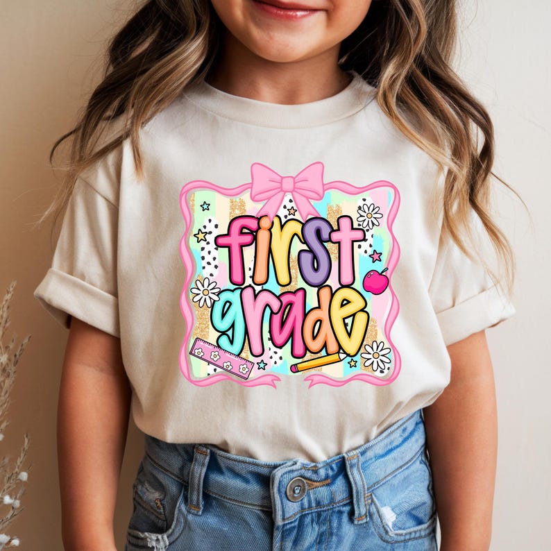 May include: A beige t-shirt featuring a colorful graphic that says "First Grade." The design includes a pink bow, stars, daisies, a ruler, a pencil, and an apple. The text is in a mix of colors, including purple, yellow, and green.