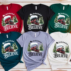 May include: A collection of short-sleeved t-shirts in various colors, including red, black, and green. Each shirt features a graphic of a train with the word "BELIEVE" in a decorative font, with a winter theme.