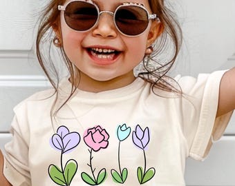 May include: A light beige t-shirt with a graphic of four colorful flowers and the text "WILD & THREE".