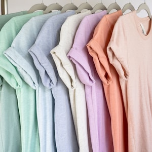 May include: A row of six plain t-shirts hanging on a rack. The shirts are in various pastel colors, including light green, light blue, beige, lavender, peach, and pink.