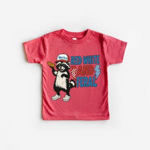 May include: Red t-shirt featuring a cartoon raccoon wearing a hat that says "'Merica" and holding a hot dog. The shirt has the words "Red White and Feral" in red and blue text. The raccoon is wearing white shoes.
