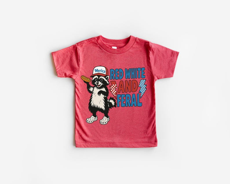 May include: Red t-shirt featuring a cartoon raccoon wearing a hat that says "'Merica" and holding a hot dog. The shirt has the words "Red White and Feral" in red and blue text. The raccoon is wearing white shoes.