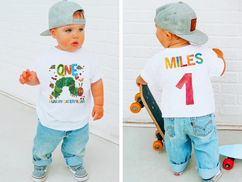 May include: A toddler wearing a white t-shirt with a colorful "ONE" and "Hungry Caterpillar" graphic, paired with blue jeans and a gray cap. The back of the shirt displays "MILES" and the number "1". The child is holding a skateboard.