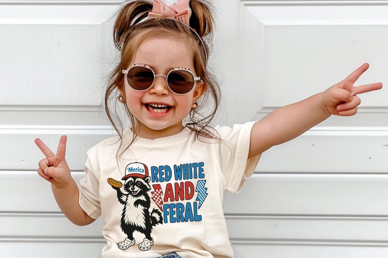 May include: A young child wearing a cream-colored t-shirt with a graphic of a raccoon holding a hot dog and the words "Red White and Feral." The child is wearing sunglasses and making a peace sign with both hands.