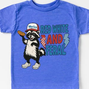 May include: A blue t-shirt featuring a cartoon raccoon wearing a hat that says "'Merica" and holding a hot dog. The shirt has the text "Red White and Feral" with lightning bolt graphics.