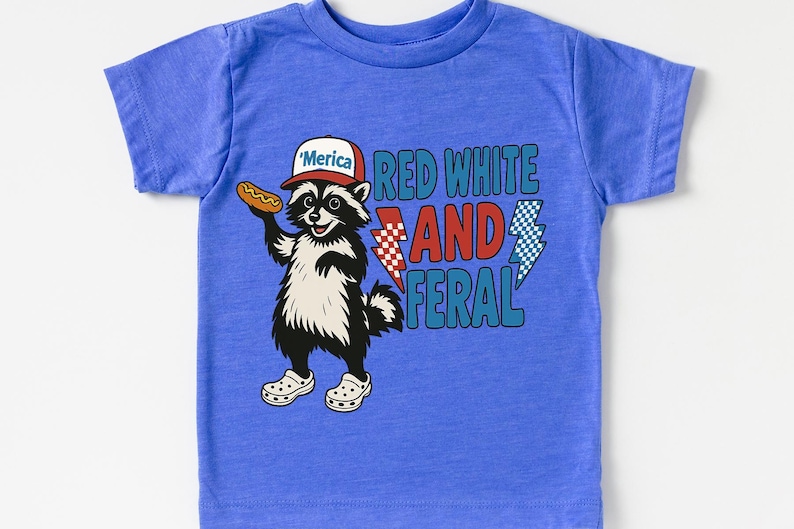 May include: A blue t-shirt featuring a cartoon raccoon wearing a hat that says "'Merica" and holding a hot dog. The shirt has the text "Red White and Feral" with lightning bolt graphics.