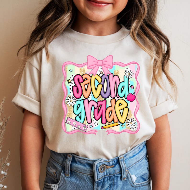 May include: A cream-colored t-shirt featuring a graphic print with the text "second grade" in colorful bubble letters. The design includes a pink bow, a ruler, a pencil, an apple, and daisies against a background of pastel stripes and gold glitter.