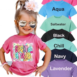 May include: A short-sleeved t-shirt in various colors including hot pink, aqua, saltwater, black, chill, navy, and lavender. The pink shirt features a graphic with the text "second grade," a bow, flowers, a ruler, and a pencil.