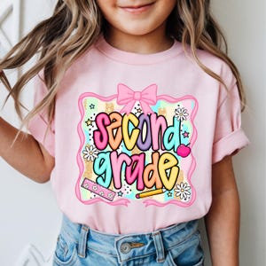 May include: A light pink t-shirt featuring a graphic print with the text second grade in colorful bubble letters. The design includes a pink bow, a ruler, a pencil, daisies, and stars against a background of pastel stripes and gold glitter.