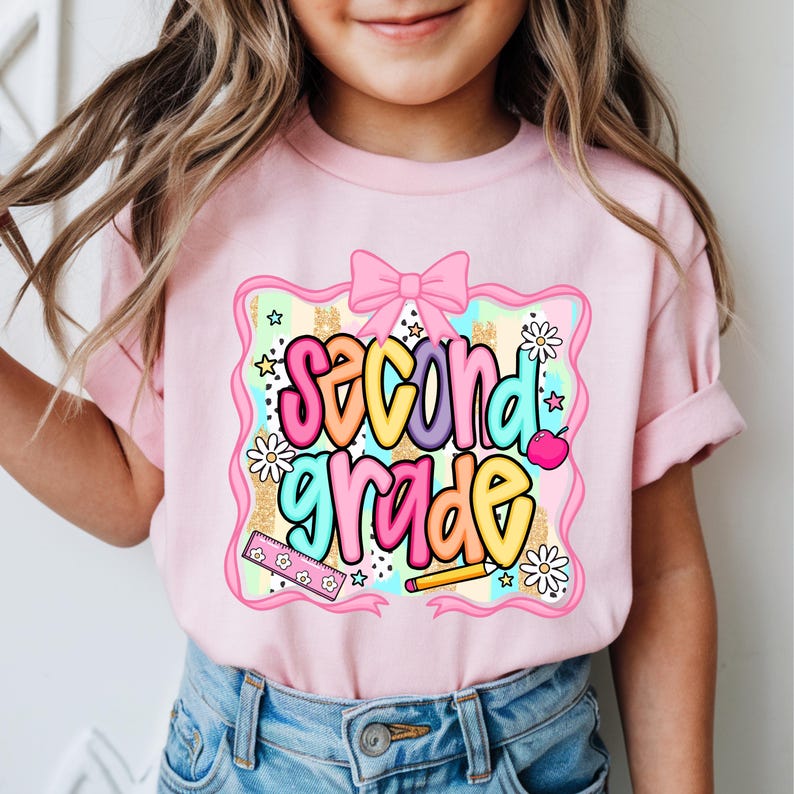 May include: A light pink t-shirt featuring a graphic print with the text second grade in colorful bubble letters. The design includes a pink bow, a ruler, a pencil, daisies, and stars against a background of pastel stripes and gold glitter.