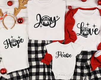 Puede incluir: Un conjunto de cuatro camisetas blancas con una escena de la natividad negra y las palabras "Hope", "Joy", "Peace" y "Love" impresas en ellas. Las camisetas están dispuestas sobre un fondo a cuadros blanco y negro con decoraciones navideñas rojas.