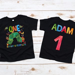 May include: Two black t-shirts with colorful designs. One shirt features a green caterpillar and the text "ONE" and "HUNGRY CATERPILLAR." The other shirt has the name "ADAM" and the number "1" in colorful letters.