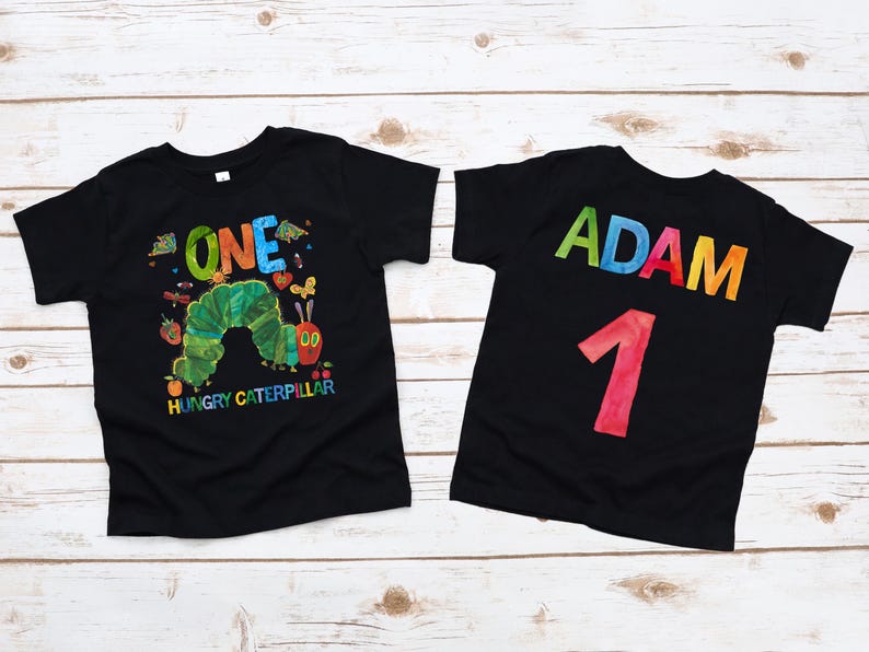 May include: Two black t-shirts with colorful designs. One shirt features a green caterpillar and the text "ONE" and "HUNGRY CATERPILLAR." The other shirt has the name "ADAM" and the number "1" in colorful letters.