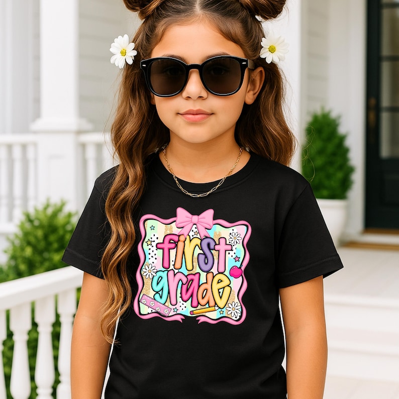 May include: A black t-shirt with a colorful graphic that says "First Grade." The graphic includes a pink bow, stars, flowers, and school supplies. The girl in the image is wearing sunglasses and a gold necklace.