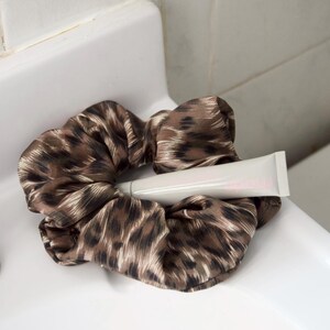 May include: A brown and black leopard print scrunchie with a white tube of product resting on top. The scrunchie is made of a silky material and is sitting on a white surface.