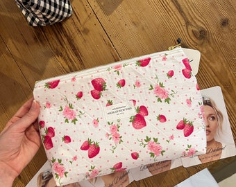 May include: White quilted makeup bag with a pink and white strawberry print. The bag has a gold zipper and the text "Rachel Dishnoit Made in New York Est. 2015" is printed on the front.