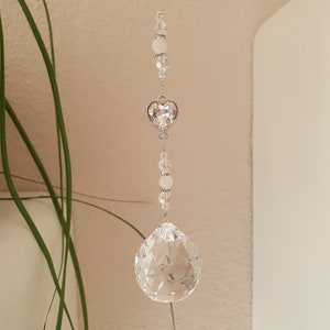 May include: A crystal suncatcher with a clear crystal ball and a silver heart charm. The suncatcher is strung with clear and white beads.