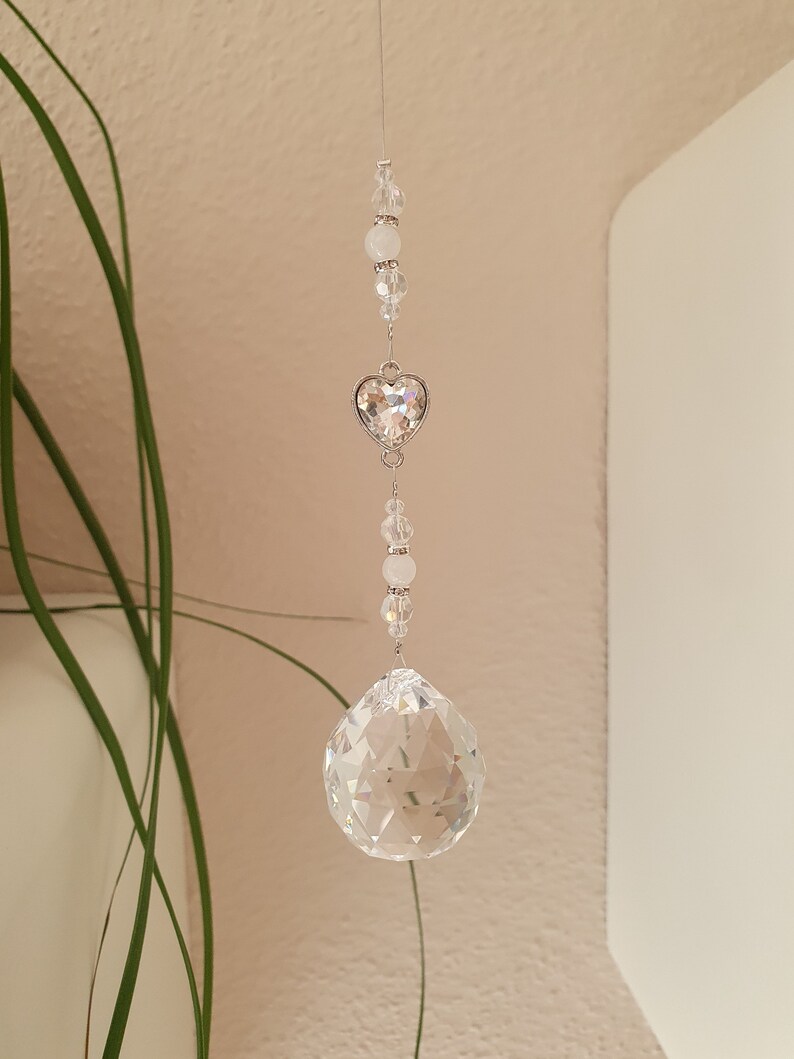 May include: A crystal suncatcher with a clear crystal ball and a silver heart charm. The suncatcher is strung with clear and white beads.