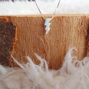 Könnte beinhalten: Eine silberne Kettenkette mit einem weißen Opal-Blitz-Anhänger. Die Kette wird auf einer Holzoberfläche mit weißem Fell präsentiert.