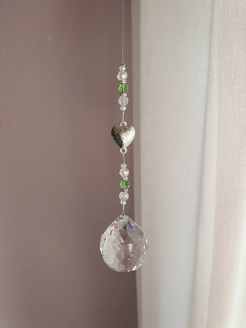 May include: A crystal suncatcher with a silver heart charm and green and clear beads hanging from a white string.