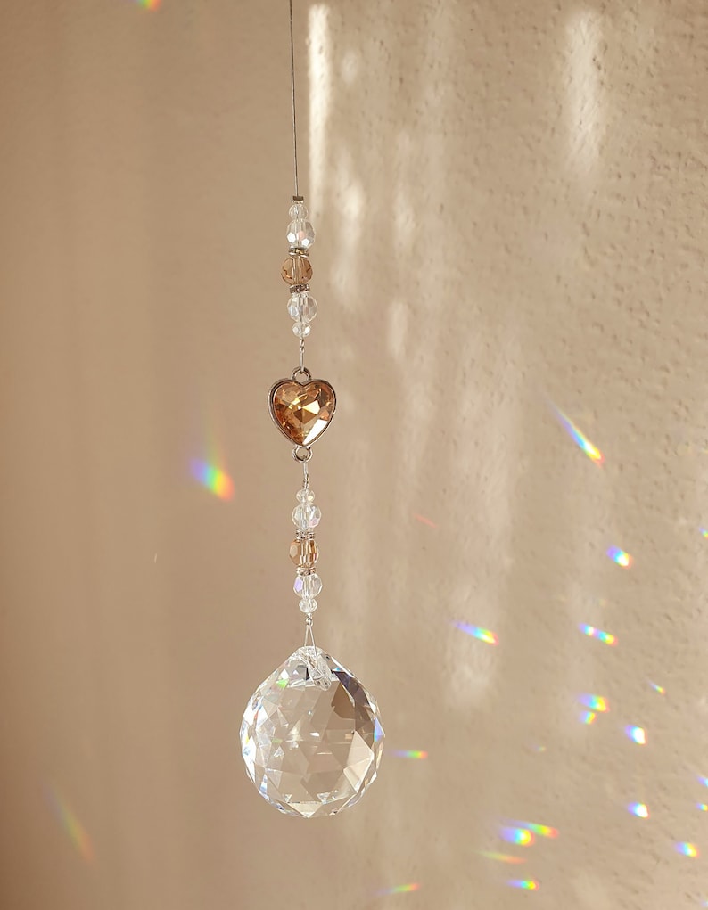 May include: A crystal suncatcher with a clear crystal ball and a heart-shaped crystal pendant. The pendant is a light brown color and the crystal ball is clear. The suncatcher is hanging from a silver string.