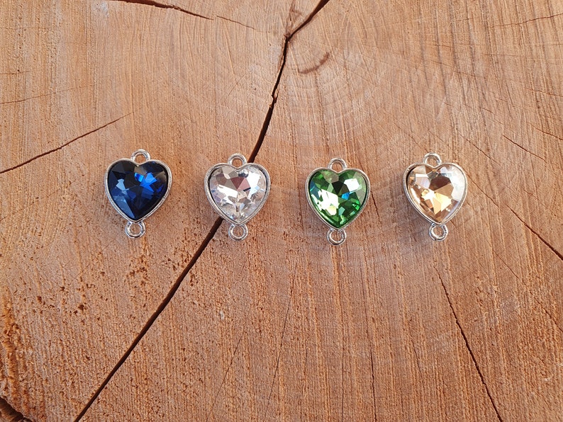 May include: Four heart-shaped charms with crystal accents in blue, clear, green, and gold. Each charm has a silver metal loop on the top and bottom.