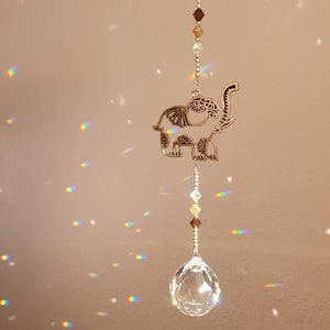 Könnte beinhalten: Ein Kristall-Sonnenfänger mit einem goldenen Elefanten-Design und klaren Kristallperlen, die an einer klaren Kristallkugel hängen. Der Sonnenfänger wirft Regenbögen an die Decke.