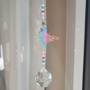 Könnte beinhalten: Ein Kristall-Sonnenfänger mit einem regenbogenfarbenen, schillernden Schmetterlingsanhänger und einer klaren Kristallkugel, die an einer silbernen Kette mit bunten Perlen hängt.