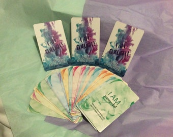 May include: A collection of affirmation cards with colorful watercolor designs. Three cards display "I AM ENOUGH!" with "Daily Confidence Affirmations." Additional cards show affirmations like "I AM resourceful." The cards are on a light green and purple background.