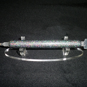 Op de afbeelding: Een glitterende, veelkleurige diamond painting pen met een doorzichtige plastic punt en een goudkleurige metalen punt. De pen wordt getoond op een heldere acryl standaard met een gebogen basis. De achtergrond is zwart.