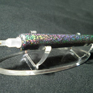 Op de afbeelding: Een zwarte glitterpen met een doorzichtige plastic punt en een gouden metalen punt, gepresenteerd op een heldere acryl standaard. De pen is bedekt met iriserende glitter, die meerdere kleuren reflecteert. De standaard heeft een gebogen basis en twee rechte steunen.
