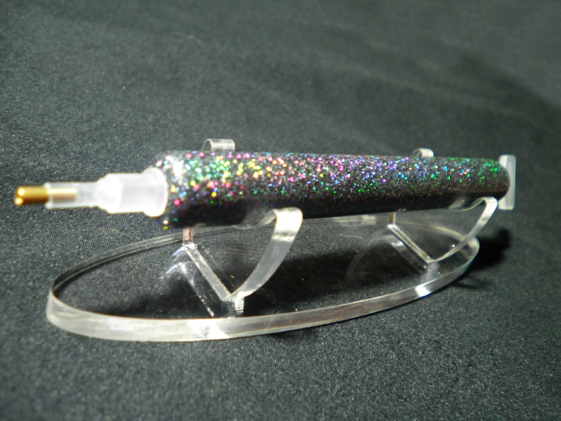 Op de afbeelding: Een zwarte glitterpen met een doorzichtige plastic punt en een gouden metalen punt, gepresenteerd op een heldere acryl standaard. De pen is bedekt met iriserende glitter, die meerdere kleuren reflecteert. De standaard heeft een gebogen basis en twee rechte steunen.