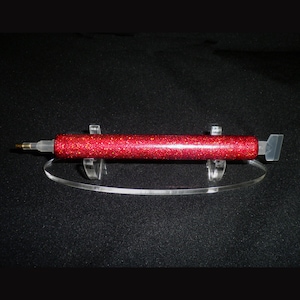 May include: A red glitter glue applicator tool with a clear tip and a silver metal scraper on the end. The tool is resting on a clear acrylic stand.