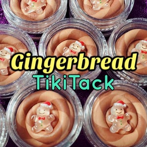 Puede incluir: Primer plano de varios frascos de vidrio pequeños llenos de un remolino de crema marrón claro, cada uno coronado con una decoración transparente en forma de hombre de jengibre con un gorro de Papá Noel. El texto "Gingerbread Tiki Tack" se muestra en amarillo y verde azulado.