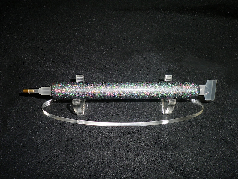 Op de afbeelding: Een glitterende, veelkleurige diamond painting pen met een doorzichtige plastic punt en een goudkleurige metalen punt. De pen wordt getoond op een heldere acryl standaard met een gebogen basis. De achtergrond is zwart.