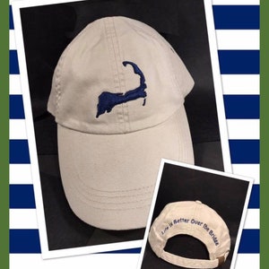 May include: A beige baseball cap with a navy blue embroidered outline of a peninsula. The cap has a curved brim and a fabric strap closure. The text "Life is Better Over the Bridge" is embroidered on the back of the cap.