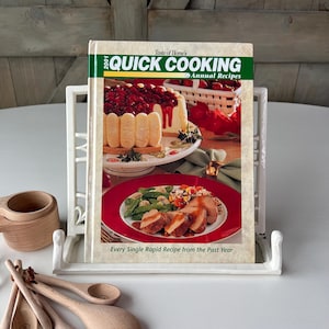 May include: A white cookbook stand holding a "Taste of Home's Quick Cooking" cookbook. The cookbook cover features images of food, including a cake and a plate of meat and vegetables. Wooden spoons and a small wooden cup are in the foreground.