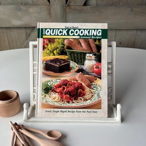 May include: A cookbook titled "Quick Cooking Annual Recipes" is displayed in a white book stand. The cover features images of prepared food. Wooden spoons and a small wooden cup are visible in the foreground. The book's title is in green and white.