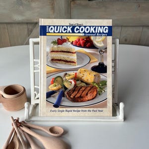 May include: A cookbook titled "Quick Cooking Annual Recipes" is displayed in a white book stand. The cover features images of food, including cake, fruit, and grilled items. Wooden measuring cups and spoons are in the foreground.