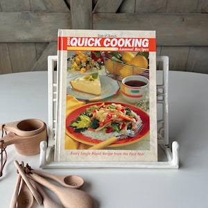 May include: A white metal cookbook stand holds a "Taste of Home's Quick Cooking" cookbook. The cookbook cover features images of food, including a slice of pie and a plate of vegetables. Wooden measuring cups and spoons are in the foreground.