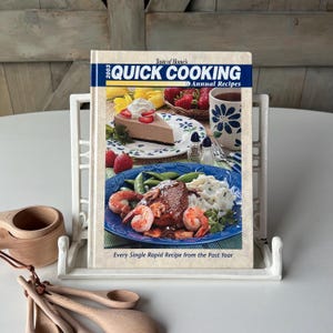May include: A cookbook titled "Taste of Home's Quick Cooking 2003 Annual Recipes" is displayed in a white metal cookbook stand. The cover features images of food, including a slice of cake, shrimp, and strawberries. Wooden measuring spoons and cups are visible.